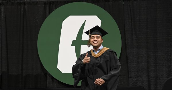 Belk College of Business MBA graduate walking across stage in front of green Charlotte logo, showing the thumbs up symbol