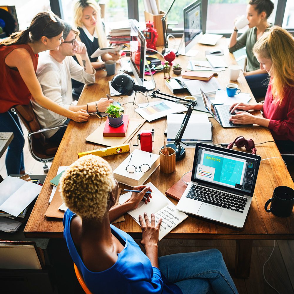 Undergraduate program in marketing, marketing concentration. Group of coworkers at a large desk working on laptops and taking notes in notebooks.  