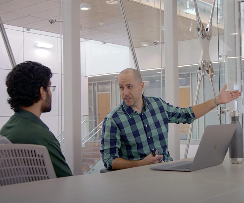 Justin Webb speaks with a student during a mentoring session. The two are seated at a table in a modern collaborative space, engaged in discussion about entrepreneurship and innovation.