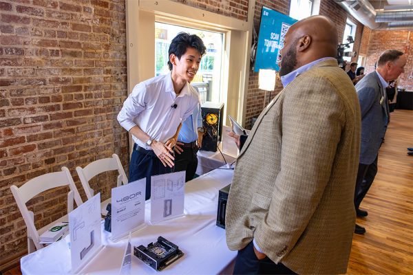 A UNC Charlotte student presents his business pitch to a visitor during The Board of Visitors convening at CO-LAB at UNC Charlotte Center City. The student gestures toward a table displaying computer hardware and diagrams, engaging in a lively discussion about his entrepreneurial concept.