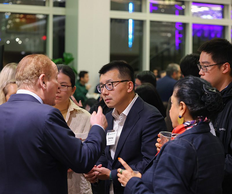 Alumnus Jindong Zhao ’16, wearing a name tag and business attire, speaks with Nobel laureate Professor Robert C. Merton following the Mathematical Finance 20th anniversary event at The Dubois Center at UNC Charlotte Center City. The group, dressed in professional and business-casual attire, is engaged in conversation in a modern indoor setting with warm lighting and a networking atmosphere.