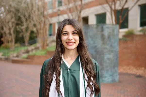 Joy Kamel, wearing a green graduation gown and white honor cords, stands smiling in front of a campus building at UNC Charlotte. Leafless trees and a cylindrical sculpture appear in the background, capturing a proud moment of achievement at the Belk College of Business.