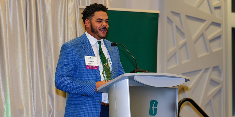 Austin McNeill ’18 in a blue jacket and green tie behind a UNC Charlotte podium with his 10 Under Ten award