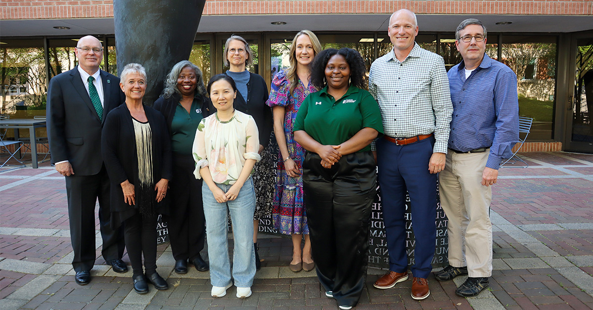 Dean Richard Buttimer stands with the 2025–26 Belk College of Business faculty and staff award winners outside the Friday Building at UNC Charlotte. The group of nine smiles together in front of the campus sculpture, representing this year’s honorees for excellence in teaching, scholarship, service, and staff achievement.