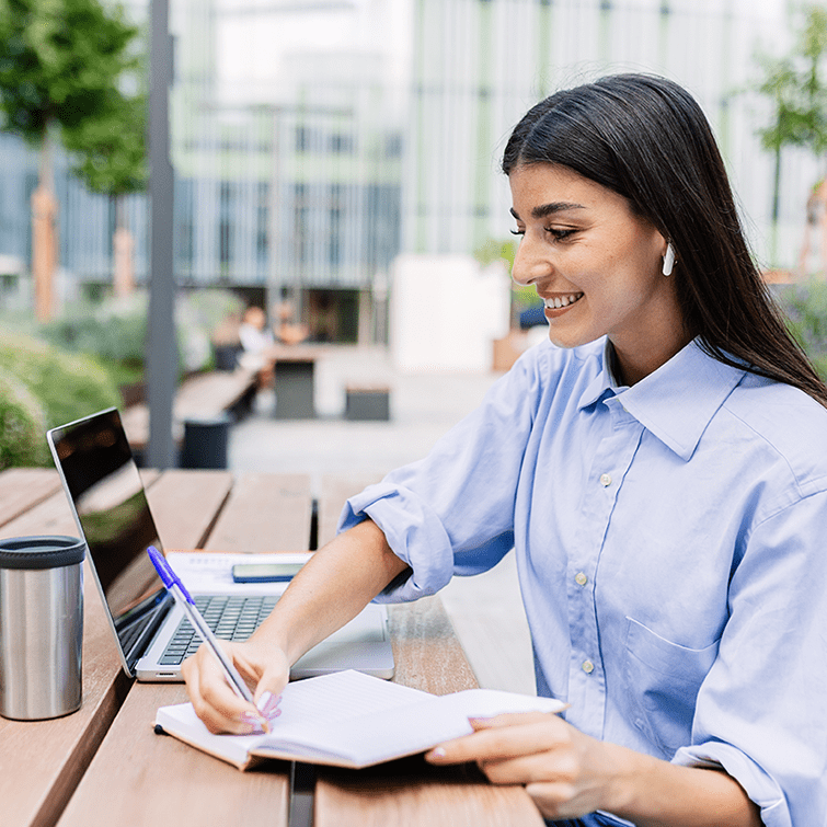 Woman sitting at an outside table with an open laptop computer, writing on a notebook