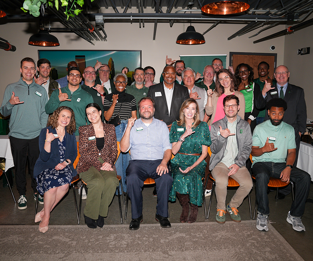 Group photo of the 2025–26 Belk College of Business Alumni Council. Members pose indoors, smiling and making the pickaxe hand gesture to show Niner pride. The group includes a mix of seated and standing participants dressed in business casual and green attire, with copper light fixtures and a Charlotte skyline photo in the background.