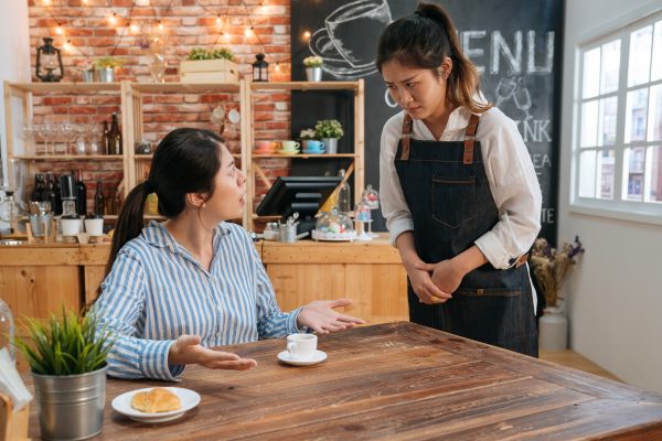 young girl waitress bowed and saying sorry while female customer complaining. coffeehouse staff woman looks at guest with concern while stand at table where client sit in cafe. bad service concept