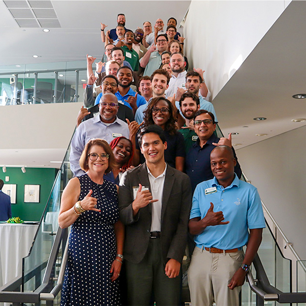 2026-2027 Alumni Council members on the second floor staircase of The Dubois Center at UNC Charlotte Center City making the pickaxe hand gesture 